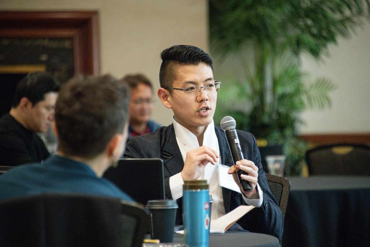 Young man in glasses speaks into a microphone during a meeting, holding notes and gesturing calmly as attendees listen in a conference room.