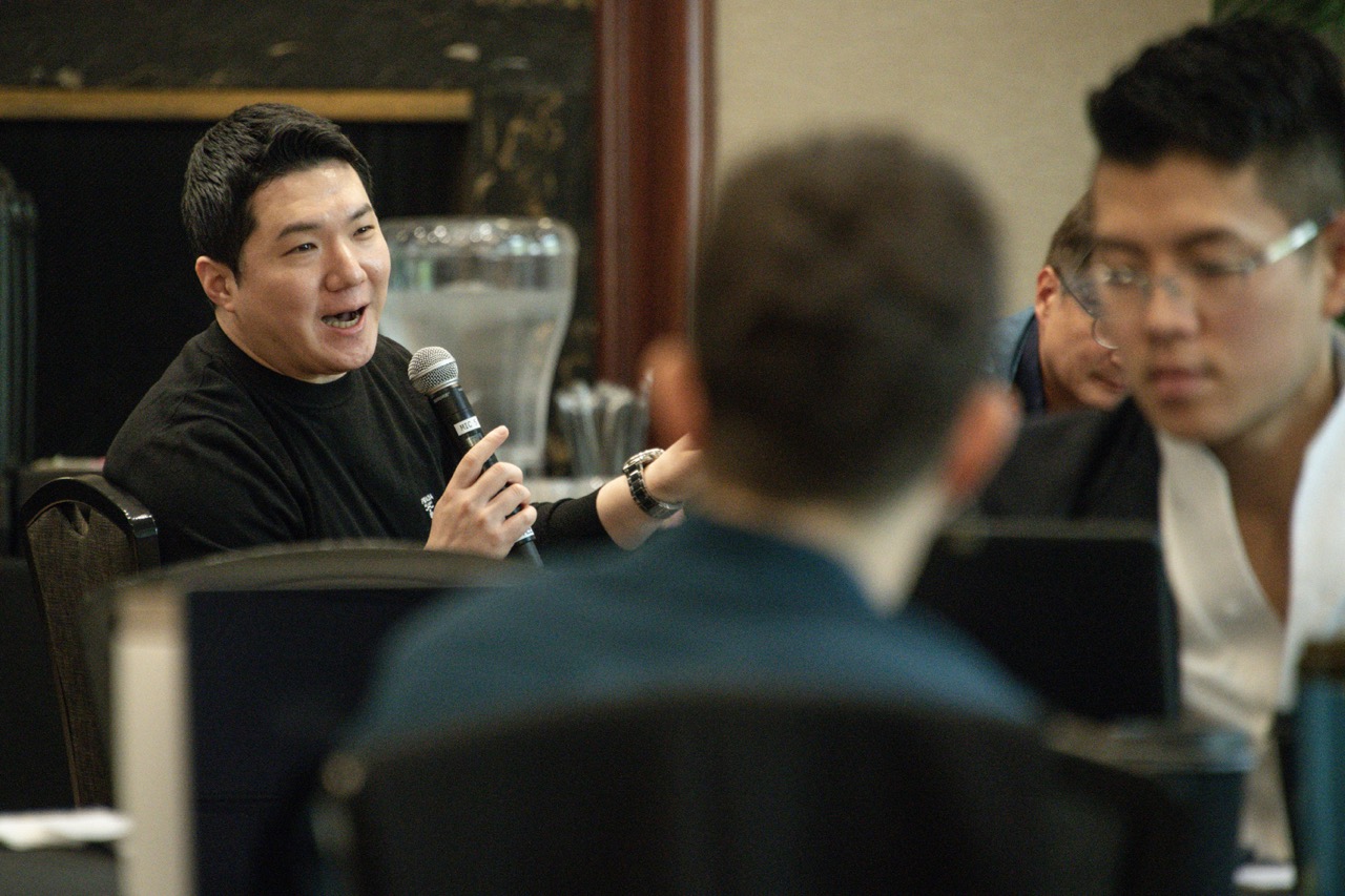 A speaker holds a microphone during a lively discussion, addressing an audience seated around tables in a warm indoor setting.
