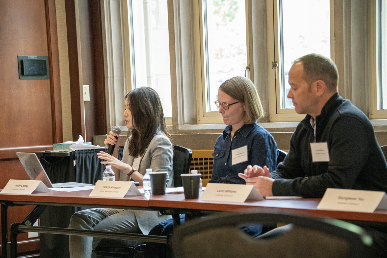 Three panelists sit at a conference table in a wood-paneled room; a woman speaks into a microphone as two colleagues listen, with laptops and coffee.