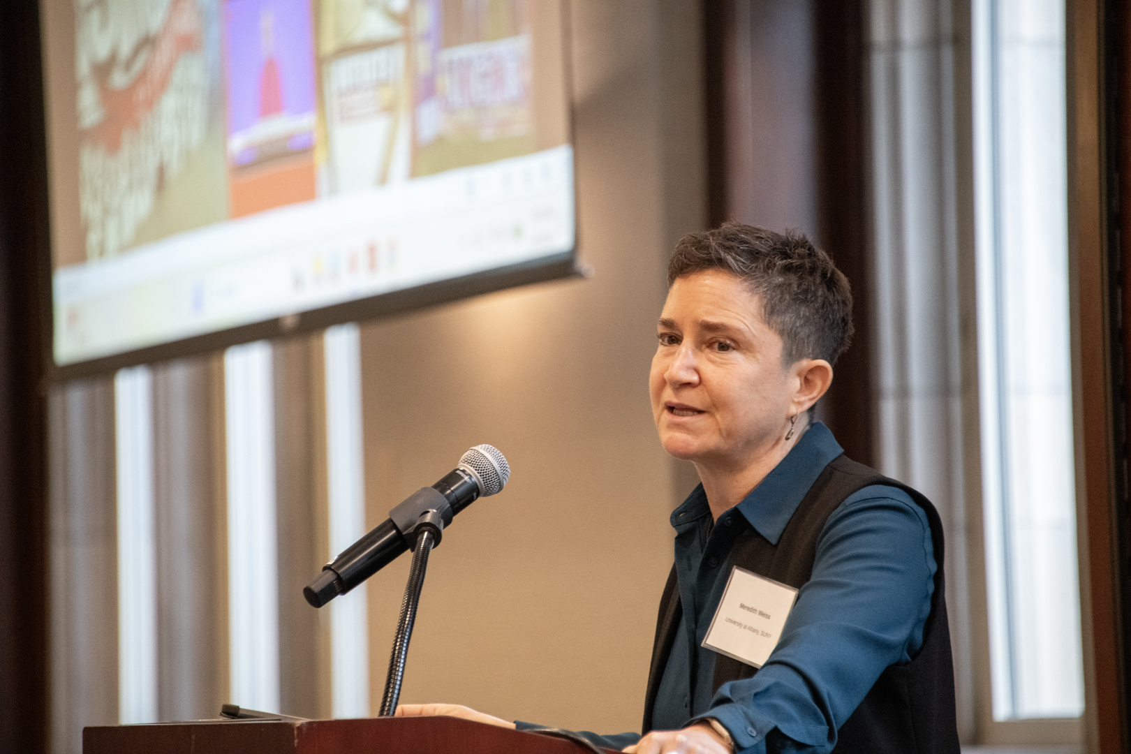 Speaker at a podium addresses an audience beside a microphone, with a presentation projected behind them, creating a focused, professional mood.