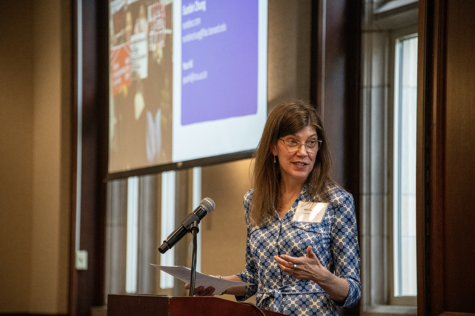 Woman in a blue patterned dress speaks at a podium beside a microphone, with a presentation slide projected behind her; professional, engaged tone.