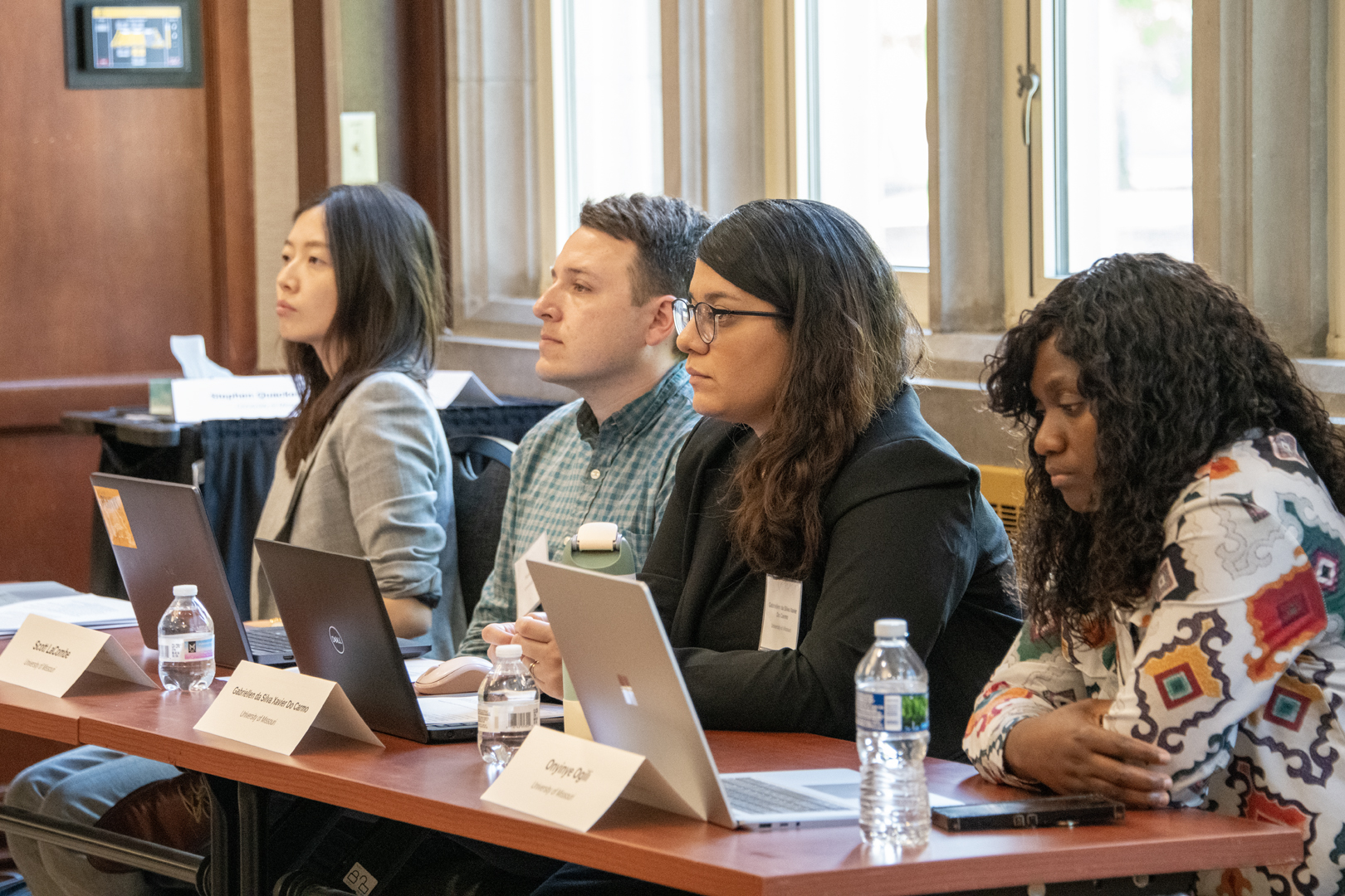 Four diverse attendees sit at a conference table with laptops and water bottles, listening attentively in a bright, professional meeting.