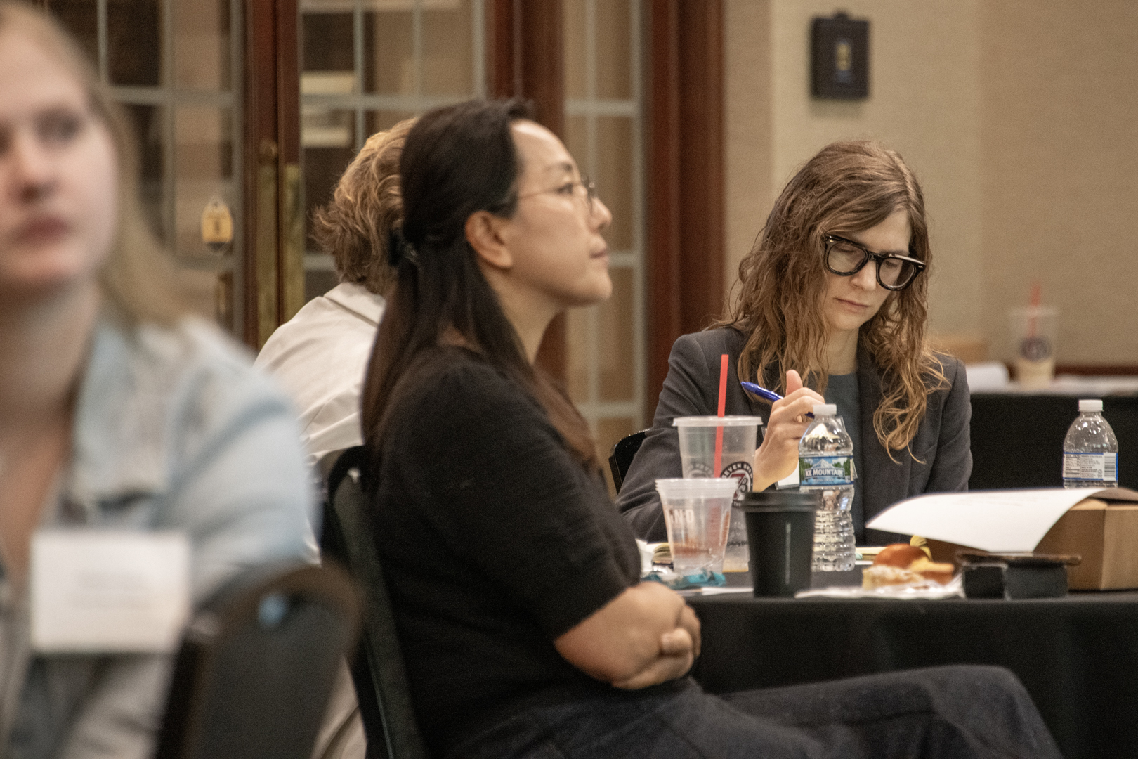 At a conference, attendees sit at a table; a woman in glasses reviews notes beside drinks and lunch, while another listens in a focused room.