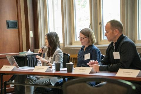 Three panelists sit at a conference table in a wood-paneled room; a woman speaks into a microphone as two colleagues listen, with laptops and coffee.
