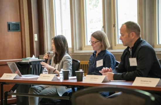 Three panelists sit at a conference table in a wood-paneled room; a woman speaks into a microphone as two colleagues listen, with laptops and coffee.